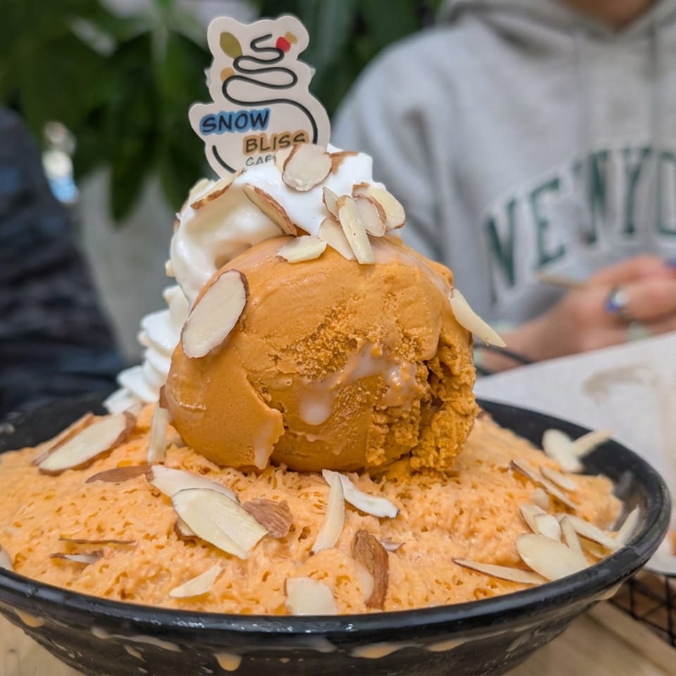 Orange shaved ice dessert topped with ice cream, whipped cream, sliced almonds, and a Snow Bliss Cafe sign.