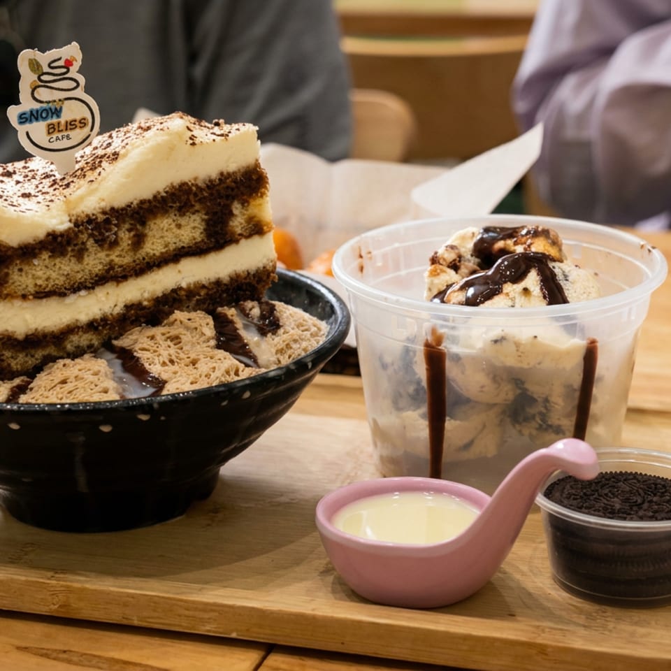 Korean shaved ice dessert topped with tiramisu cake, cereal pieces, chocolate sauce, cream, and crushed cookies on a wooden tray.